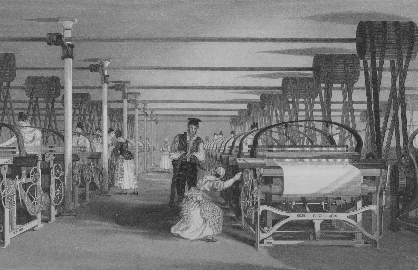 An engraving of a long mill floor lined with power looms, with belts running up to shafts in the ceiling, women tending the machines and a man in a top hat watching.