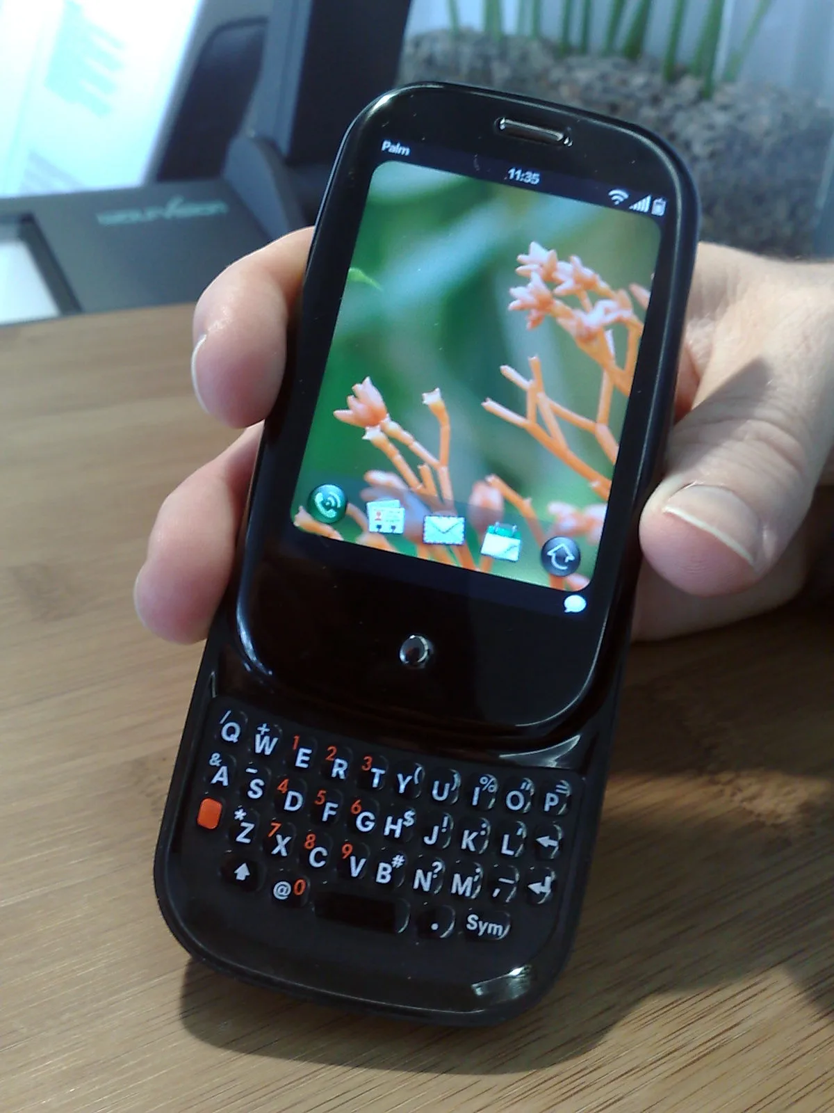 A hand holding a Palm Pre with its keyboard open, the screen showing a photo wallpaper and a row of app icons.