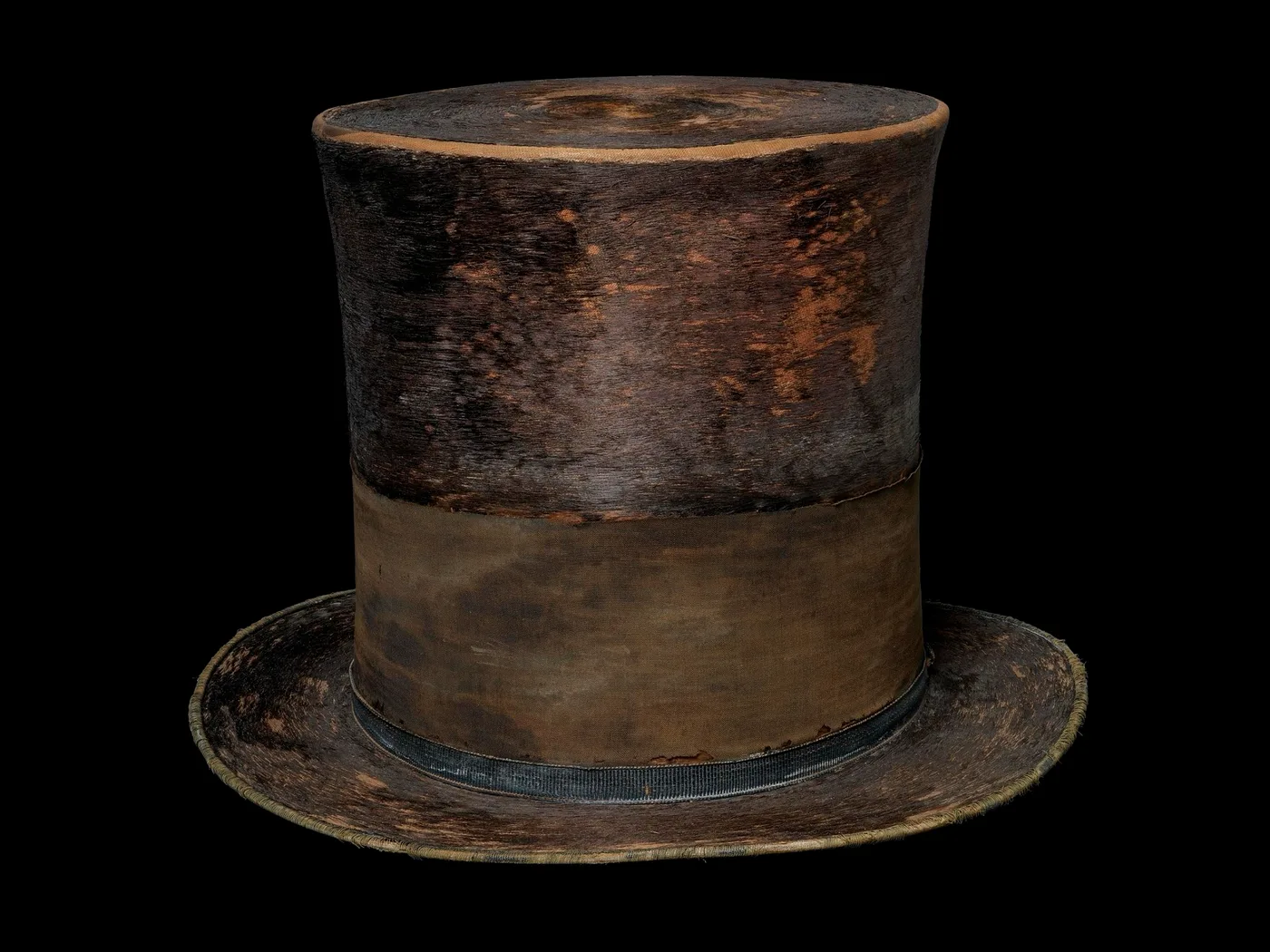 A worn black silk top hat with a tall crown and a wide black mourning band, photographed against black.