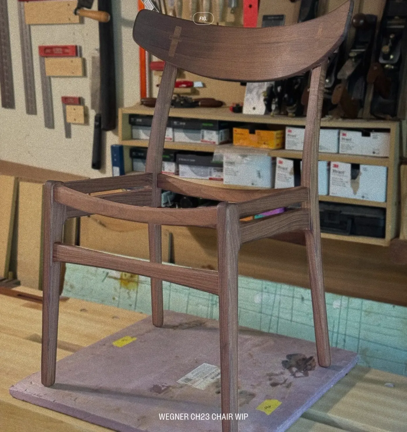 An unfinished walnut Wegner CH23 chair frame, without its seat, on a workbench in front of a wall of tools.