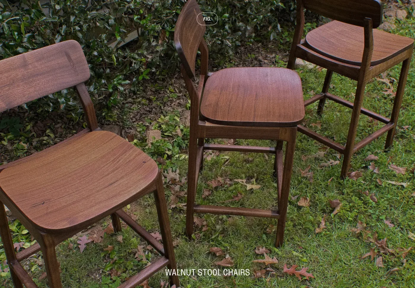 Three walnut counter stools with curved backs and scooped seats, standing on grass among fallen leaves.