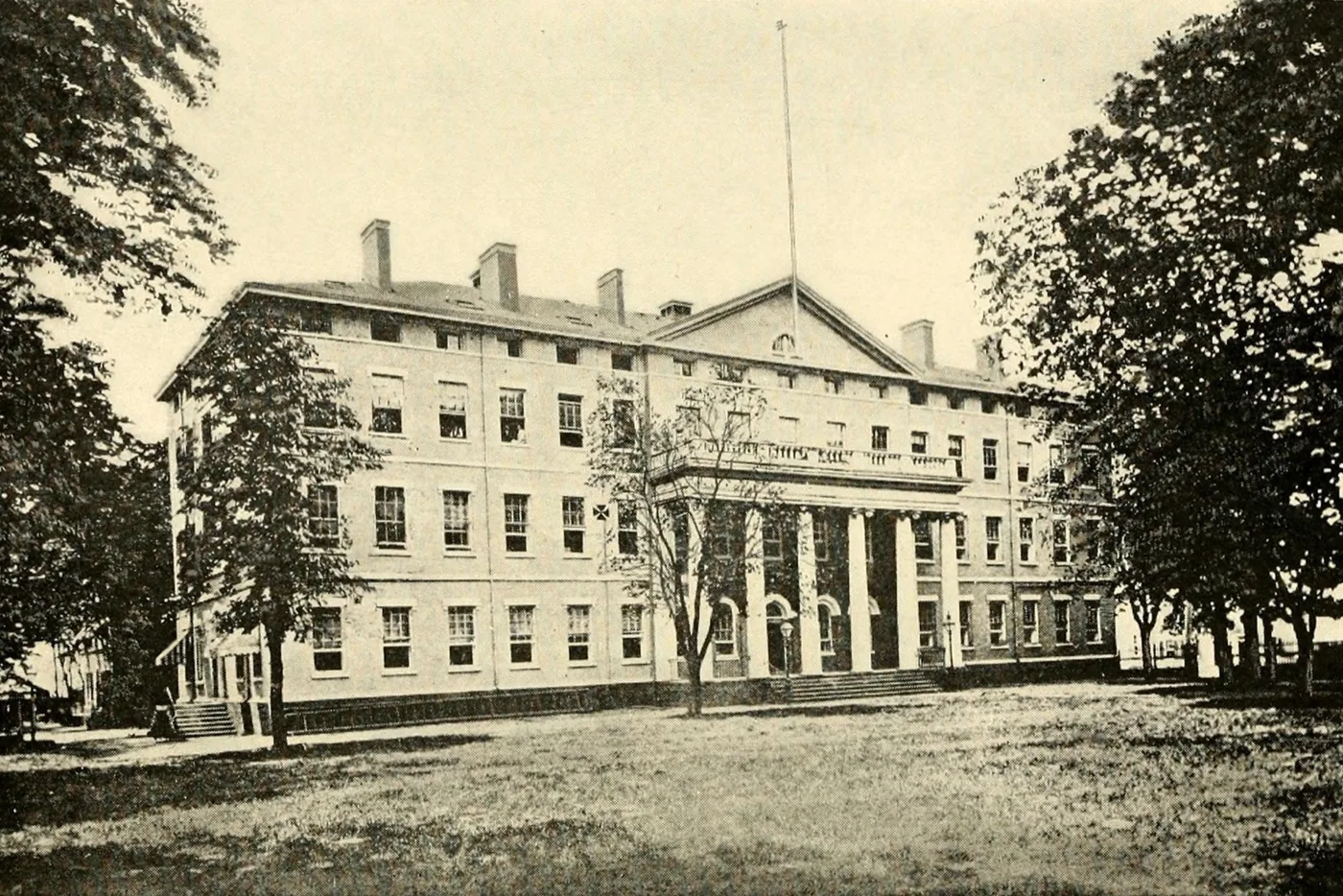 A photograph of a plain four-story building with a columned portico and a flagpole, set back behind a lawn and tall trees.